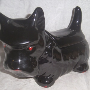 May include: Black ceramic cookie jar shaped like a Scottish terrier dog. The dog has red painted eyes and a red painted nose.