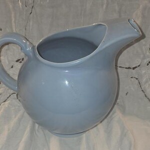 May include: A light blue ceramic pitcher with a rounded body and a curved handle. The pitcher has a spout for pouring and is likely used for serving beverages. The pitcher is approximately 8 inches tall.