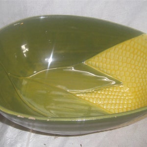 May include: A green and yellow ceramic bowl with a corn cob design. The bowl is oval shaped and has a glossy finish.