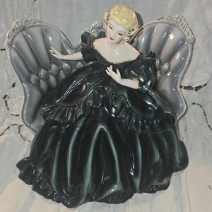 May include: A vintage porcelain figurine of a woman in a dark green dress, seated in a gray chair. The figurine has blonde hair and is adorned with gold accents. The dress features ruffled details.