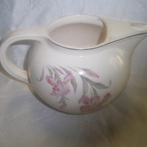 May include: A white ceramic pitcher with a floral design featuring pink and purple flowers. The pitcher has a handle and a spout.