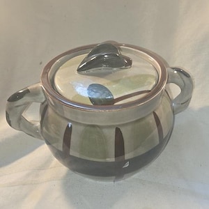 May include: A ceramic sugar bowl with a lid and two handles. The bowl is painted with a mix of green, brown, and gray colors. The lid has a black handle. The bowl is round and has a glossy finish.