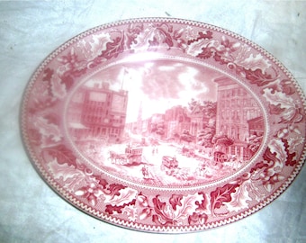 May include: A pink oval ceramic plate with a detailed design of a city scene with buildings, trees, and horse-drawn carriages. The plate has a decorative border of oak leaves and acorns.