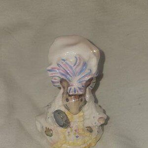 May include: A ceramic figurine of a mouse wearing a white bonnet with blue and pink accents. The mouse is dressed in a white dress with yellow and blue floral details, holding a small, gray tambourine.