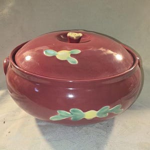 May include: A vintage red casserole dish with a lid. The dish has a floral design with green leaves and yellow flowers.