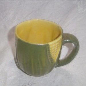 May include: A green and yellow ceramic mug shaped like an ear of corn. The mug has a handle and a yellow interior.