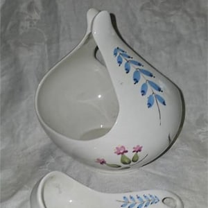 May include: White ceramic gravy boat with a blue and pink floral design and a matching ladle. The gravy boat has a unique shape with a handle on the side.