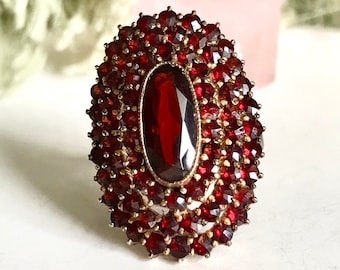 Op de afbeelding: Een vintage ring met een grote, ovale granaat omringd door een halo van kleinere granaten. De ring is gemaakt van goud en heeft een delicaat, ingewikkeld ontwerp.