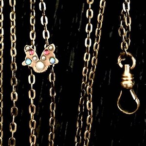 May include: A gold chain necklace with a small pendant featuring a cluster of colourful gemstones. The pendant is shaped like a bird with its wings spread.
