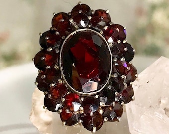 Op de afbeelding: Een vintage ring met een grote, ovale granaat omringd door kleinere granaten. De ring is gemaakt van zilver en heeft een delicaat, bloemig ontwerp.