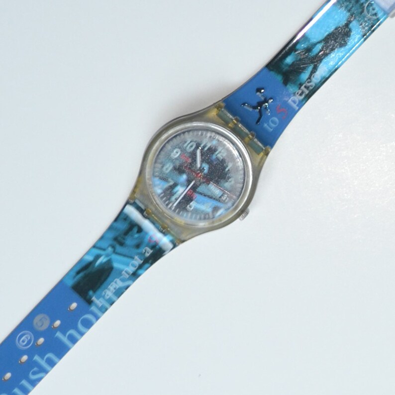 Puede incluir: Un reloj Swatch azul y blanco con una caja de pl&aacute;stico transparente. La esfera del reloj tiene n&uacute;meros y manecillas negros. La correa tiene un fondo azul con una imagen en blanco y negro de una persona corriendo. El texto "to person" est&aacute; impreso en la correa.