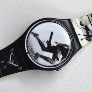 Puede incluir: Reloj de pulsera blanco y negro con una banda negra y una esfera redonda. La esfera presenta una imagen en blanco y negro de una persona corriendo. La banda tiene una serie de im&aacute;genes en blanco y negro de personas haciendo diferentes deportes.