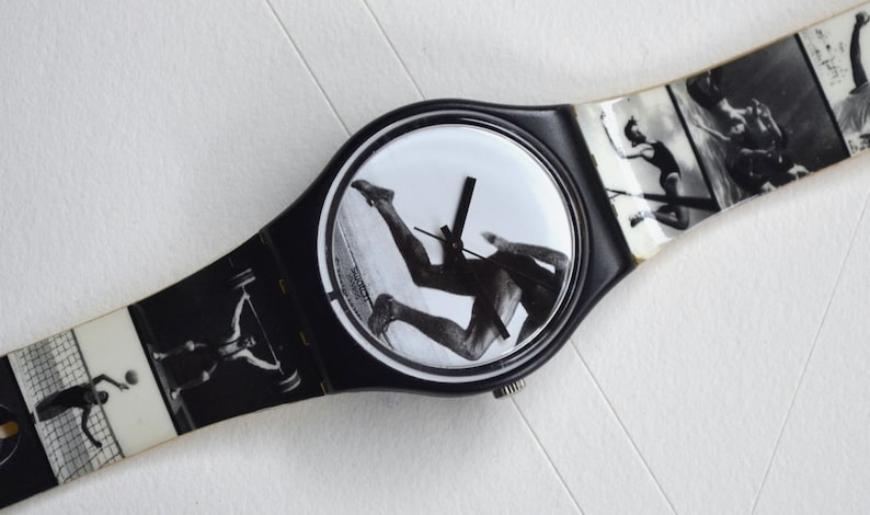 Puede incluir: Reloj de pulsera blanco y negro con una banda negra y una esfera redonda. La esfera presenta una imagen en blanco y negro de una persona corriendo. La banda tiene una serie de im&aacute;genes en blanco y negro de personas haciendo diferentes deportes.