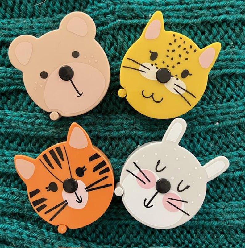 May include: Four colorful measuring tapes shaped like animal faces. The tapes are yellow, orange, pink, and gray with black eyes and white accents.