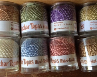 May include: Eight spools of Anchor Topas crochet thread in various colors, including beige, light green, purple, gold, ivory, blue, pink, and brown. Each spool is labeled with the brand name and the text "Topas Häkel-Becher".