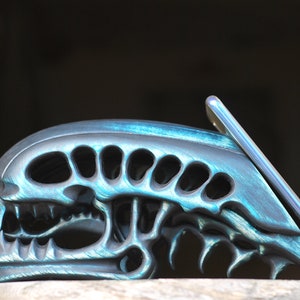 May include: A teal and black alien skull phone stand. The stand is made of a textured material and has a detailed design. The phone is not included.