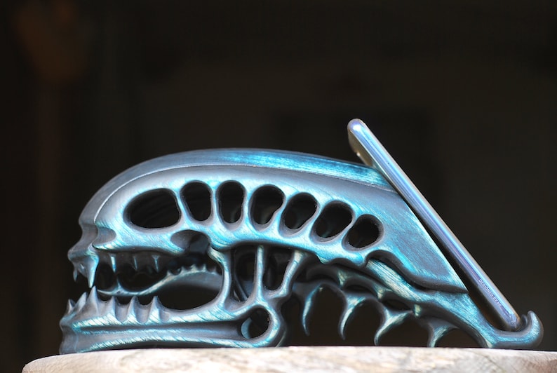 May include: A teal and black alien skull phone stand. The stand is made of a textured material and has a detailed design. The phone is not included.