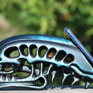 May include: A black and teal alien skull phone stand. The stand is made of wood and has a detailed design. A smartphone is resting in the stand.