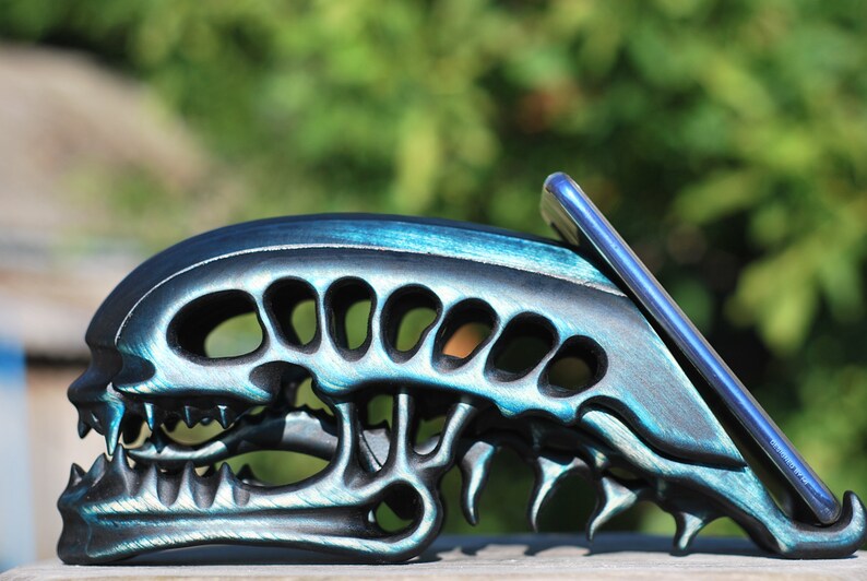 May include: A black and teal alien skull phone stand. The stand is made of wood and has a detailed design. A smartphone is resting in the stand.