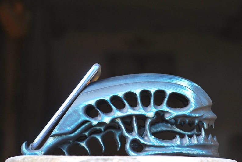 May include: A teal blue phone stand shaped like an alien head with a phone resting in the stand.