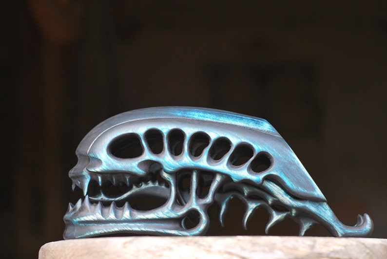 May include: A black and blue alien skull sculpture with a detailed design. The skull has a large mouth with sharp teeth and a series of holes on the top of the head.