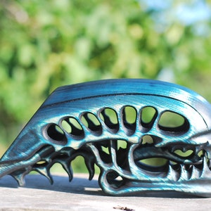May include: A teal and black alien skull sculpture with a detailed design. The skull is made of a smooth, hard material and has a glossy finish.
