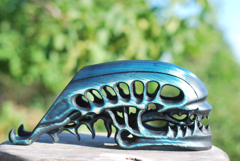May include: A teal and black alien skull sculpture with a detailed design. The skull is made of a smooth, hard material and has a glossy finish.