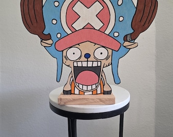 May include: A wooden cutout of the character Tony Tony Chopper from One Piece, featuring a blue hat with white dots, a pink brim, and a large white X. The character has wide eyes, an open mouth, and brown antlers, mounted on a rectangular base.