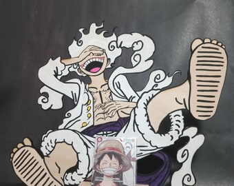 May include: A white and black cartoon character with white hair and a purple sash, holding a trading card of Monkey D. Luffy from the anime One Piece. The card is on a wooden stand.