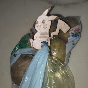 May include: A wooden Pikachu-shaped decoration is attached to a blue plastic bag. The bag is tied around a clear plastic bag containing potatoes. The potatoes are visible through the clear plastic.