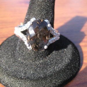 May include: A silver ring with a large, faceted, oval-shaped smoky quartz gemstone. The ring features a split shank design with small, clear accent stones. The ring is displayed on a black ring holder.