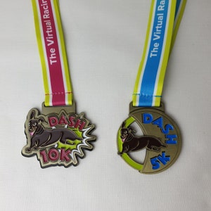 May include: Two gold-colored medals with a dachshund design. The medals are attached to ribbons. The 10K medal has a pink and yellow ribbon with the text "The Virtual Racing Dash 10K". The 5K medal has a blue and yellow ribbon with the text "The Virtual Racing Dash 5K".
