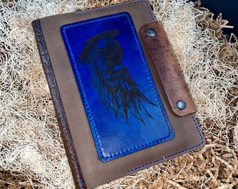May include: A brown leather journal with a blue leather flap featuring a black silhouette of a bird with wings spread. The flap has a stitched border and a brown leather strap with a metal button.