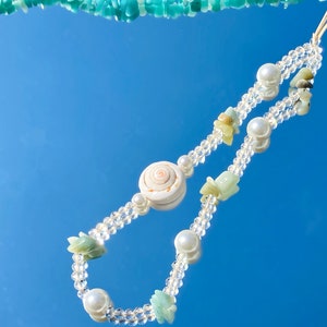 Può includere: Una delicata collana con un pendente centrale a conchiglia, perline di cristallo trasparente, perle finte e piccoli accenti di conchiglia verde chiaro. La collana è esposta su una superficie blu riflettente, creando un'estetica da spiaggia.