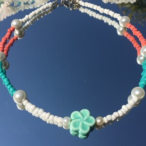 May include: A beaded necklace with white, coral, and turquoise beads, and faux pearls. The necklace has a silver clasp and a central turquoise flower charm. The necklace is displayed on a reflective surface.
