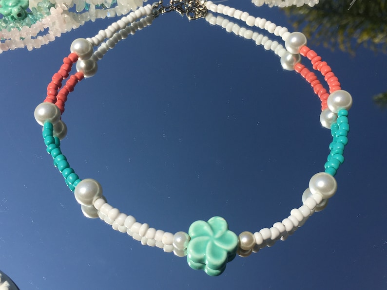 May include: A beaded necklace with white, coral, and turquoise beads, and faux pearls. The necklace has a silver clasp and a central turquoise flower charm. The necklace is displayed on a reflective surface.