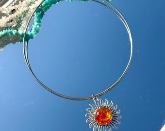 May include: A silver choker necklace with a sun pendant. The sun pendant has a silver frame with a red and yellow center. The necklace is displayed against a blue background.