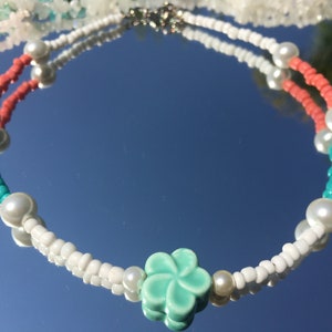 May include: A beaded necklace with white, coral, and turquoise beads, faux pearls, and a light green flower pendant. The necklace is displayed on a reflective surface.