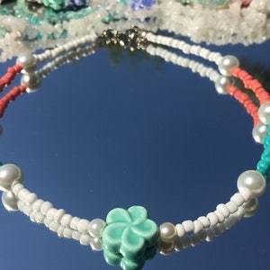 May include: A beaded necklace with white, coral, and turquoise beads, and faux pearls. A turquoise flower charm is at the centre. The necklace is displayed on a reflective surface. The necklace's design is reflected in the surface.