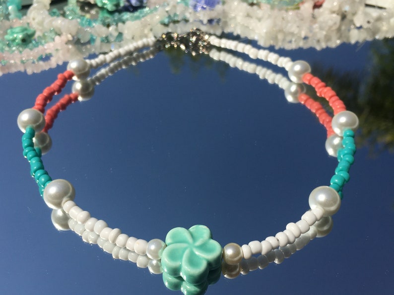 May include: A beaded necklace with white, coral, and turquoise beads, and faux pearls. A turquoise flower charm is at the centre. The necklace is displayed on a reflective surface. The necklace's design is reflected in the surface.