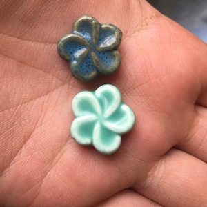 May include: Two ceramic flower beads. One is a blue-green colour with a speckled texture, and the other is a light turquoise colour. The beads are in the shape of a five-petal flower.
