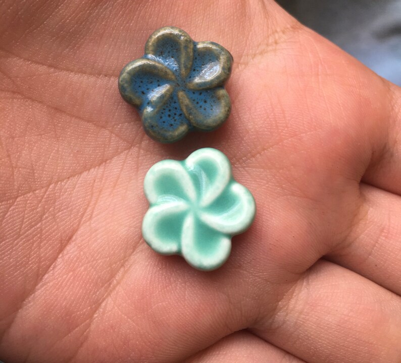 May include: Two ceramic flower beads. One is a blue-green colour with a speckled texture, and the other is a light turquoise colour. The beads are in the shape of a five-petal flower.