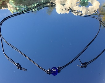 Pode incluir: Um colar de cordão preto com um amuleto azul de olho turco. O colar é amarrado com um nó na ponta.
