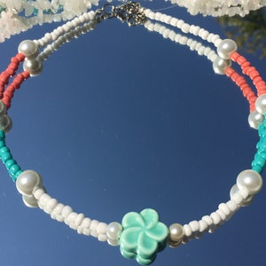 May include: A colourful necklace with white, coral, and turquoise beads, and faux pearls. The necklace has a light green flower pendant and a silver clasp. This piece of jewellery is ideal for adding a splash of colour to any outfit.