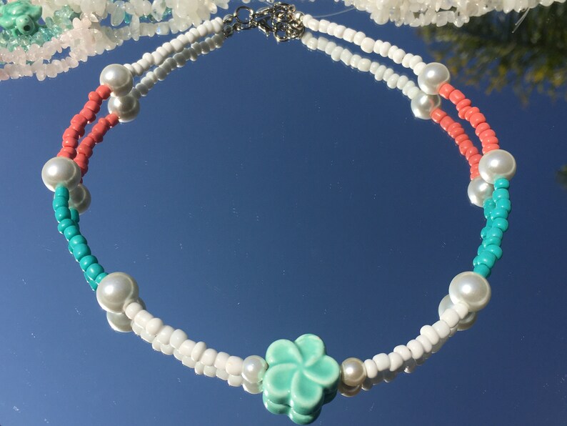 May include: A colourful necklace with white, coral, and turquoise beads, and faux pearls. The necklace has a light green flower pendant and a silver clasp. This piece of jewellery is ideal for adding a splash of colour to any outfit.