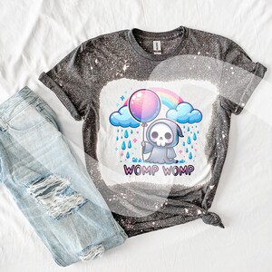 以下が含まれることがあります： 漂白されたデザインが施された黒いTシャツ。漫画の死神が虹の上にピンクの風船を持っている。死神の下に「WOMP WOMP」という文字が印刷されている。