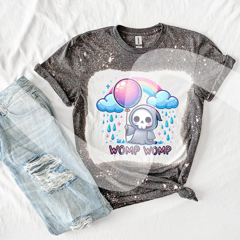 以下が含まれることがあります： 漂白されたデザインが施された黒いTシャツ。漫画の死神が虹の上にピンクの風船を持っている。死神の下に「WOMP WOMP」という文字が印刷されている。