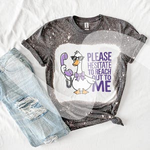 May include: A black and white bleached t-shirt featuring a cartoon goose wearing sunglasses and holding a purple phone. The text "Please hesitate to reach out to me" is printed on the shirt.