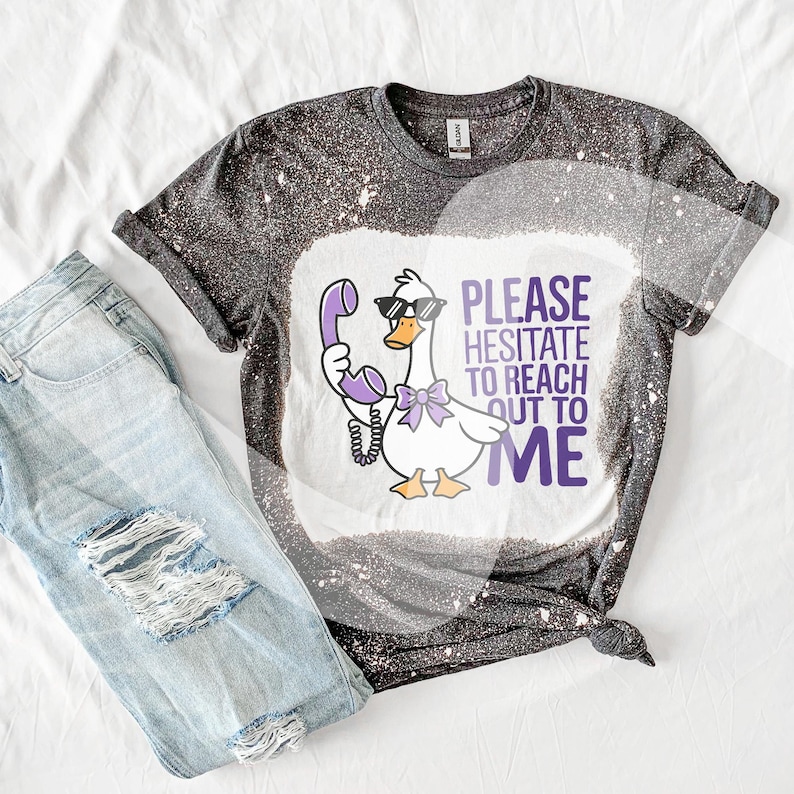 May include: A black and white bleached t-shirt featuring a cartoon goose wearing sunglasses and holding a purple phone. The text "Please hesitate to reach out to me" is printed on the shirt.