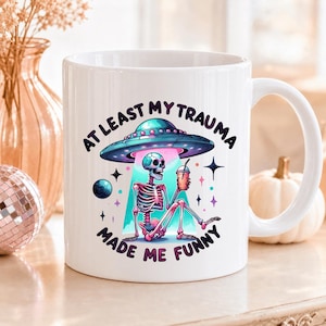 Peut inclure: Un mug en céramique blanche orné d'un graphisme coloré représentant un squelette assis sous un OVNI tenant une boisson. Le texte "AT LEAST MY TRAUMA MADE ME FUNNY" est imprimé en lettres noires incurvées au-dessus et en dessous de l'illustration.